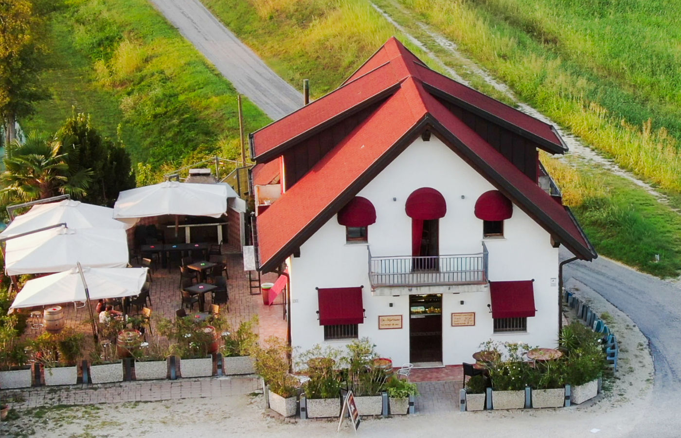 La casa dell'osteria vista dall'alto: tetto di tegole rosse, tende rosse alle finestre, il dehors con gli ombrelloni e i campi attorno